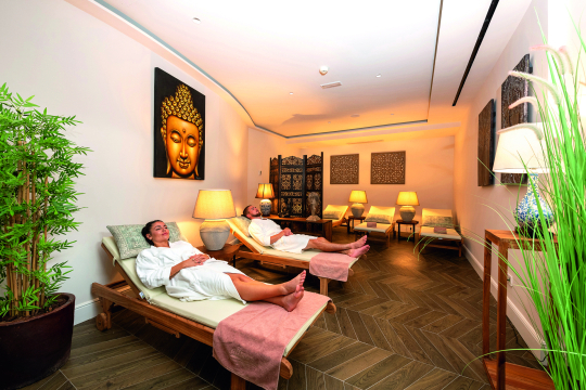 Mann und Frau entspannen im Bademantel auf einer Holzliege in einem Wellnessbereich mit Asiatischer Dekoration an den Wänden